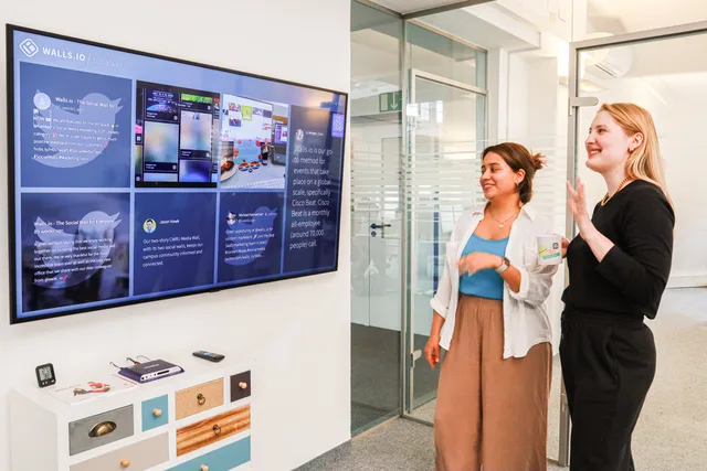 Two women in an office environment interact with a large digital display showcasing social media content. Medieplanlægning: kanaler, budget og dokumentation